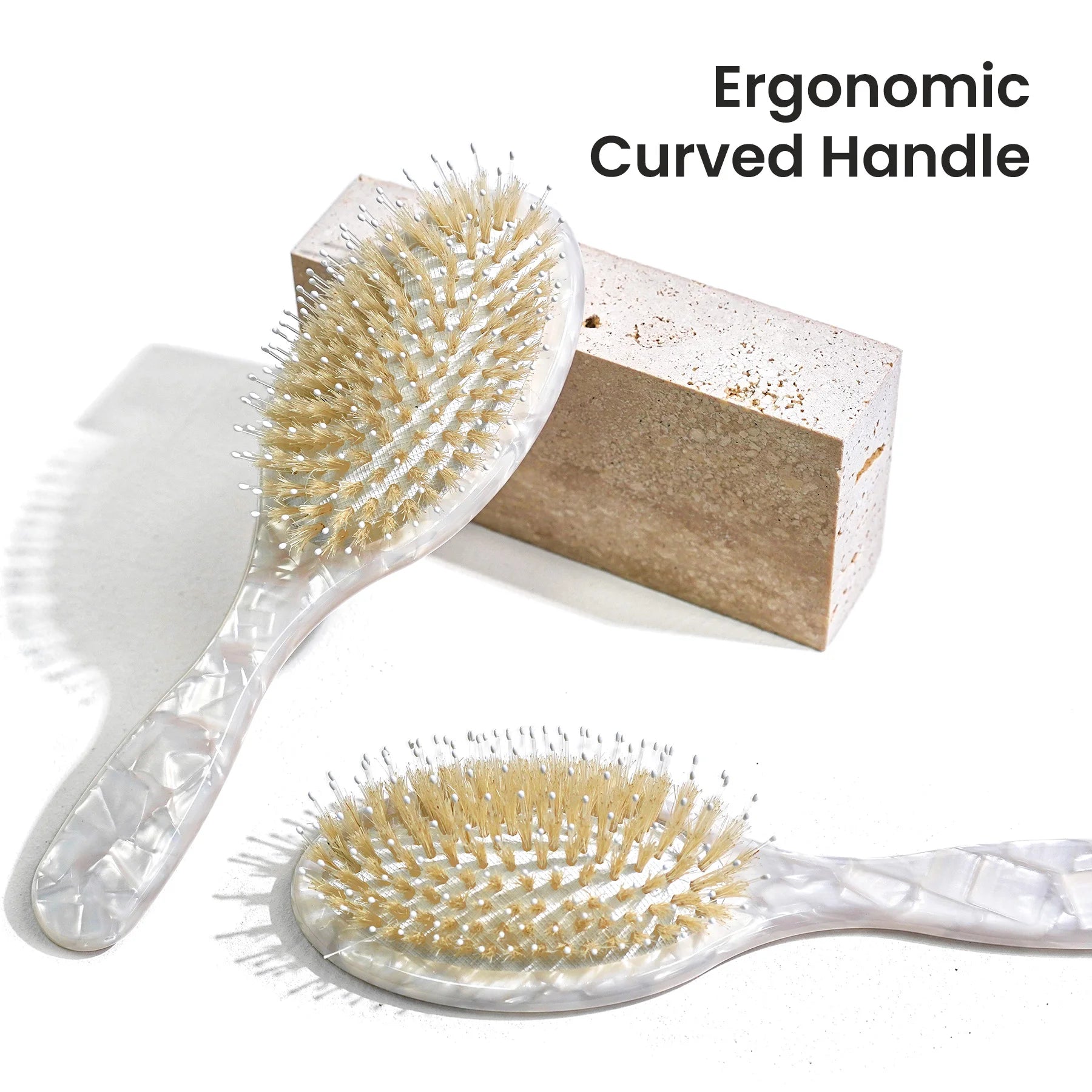 Two oval hairbrushes with curved translucent handles and beige bristles, shown on a white background with one upright behind a textured block and one lying flat. Text in the top right reads 'Ergonomic Curved Handle'.