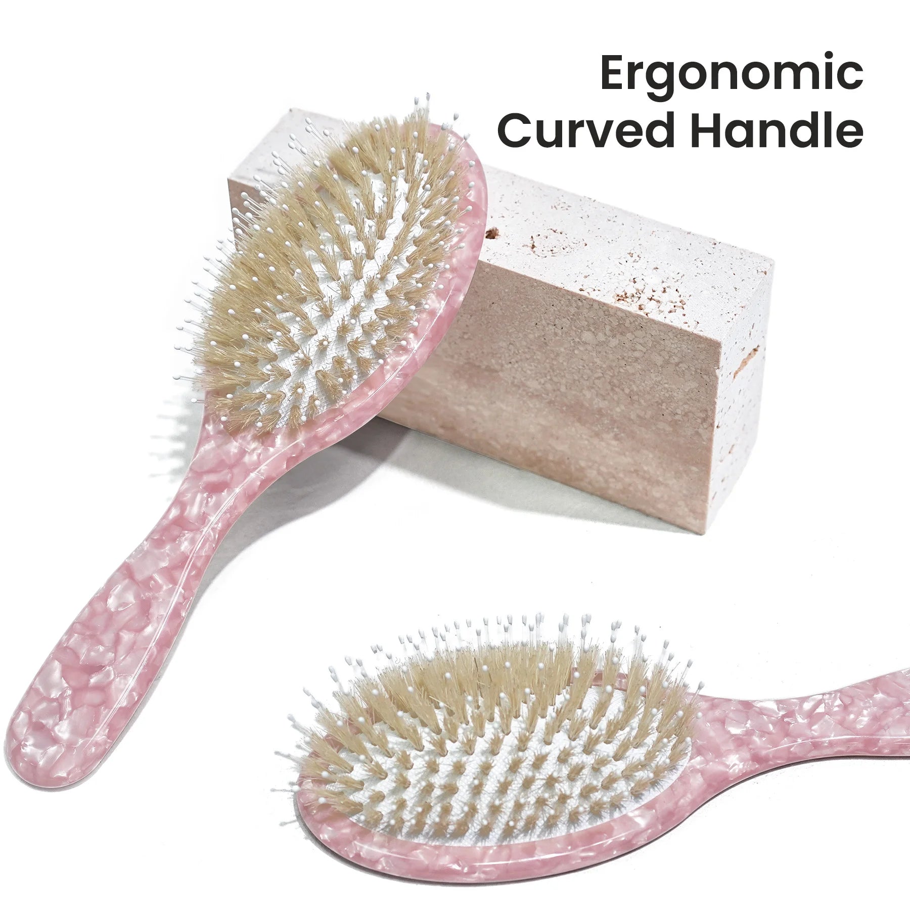 Two pink marbled hairbrushes with beige and white bristles on a white background, with one brush resting against a rectangular block and the other shown partially cropped below. Text in the top right reads 'Ergonomic Curved Handle'.